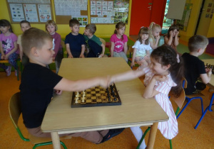 Chłopiec z dziewczynką podają sobie ręce, rozpoczynają grę w szachy.